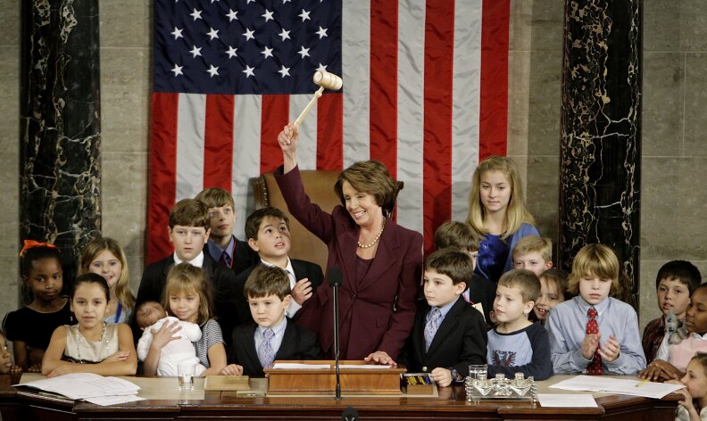 A woman in a maroon jacket holds a gavel high above her head in front of an American flag, standing at a lectern. She is surrounded by 20 young children.