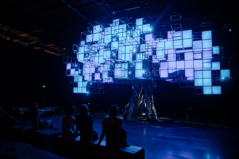 People sit in a dimly illuminated space, on wood benches, looking up at a faux tree. It's made from a real tree's trunk, with digital colored screens in square pixelated shapes creating a faux canopy. The panels at the time of this image are illuminated in light blue and purple.
