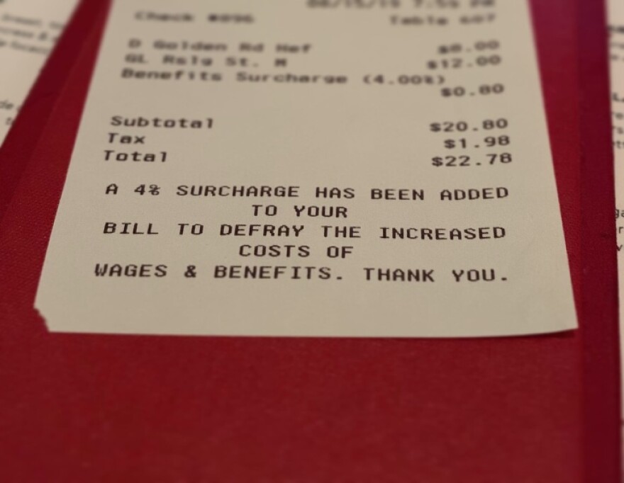 Close-up of a restaurant bill, with the words, "a 4% surcharge has been added to your bill to defray the increased costs of wages & benefits. Thank you," printed at the bottom.