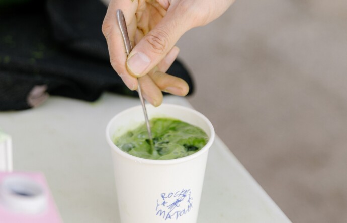 A light skinned hand hold a metal spoon as it mixes a green liquid in a paper white cup
