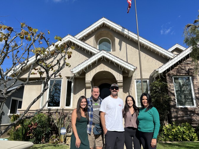 A family comprising of three females and two males stands in front of a home painted cream. The house is decorated with lights. 