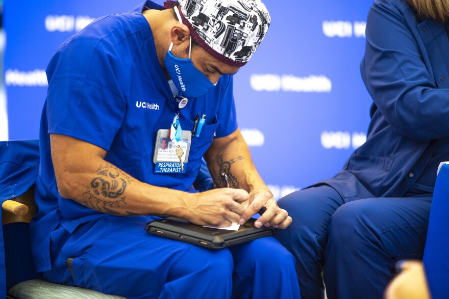 A man in blue nursing scrubs fills out a form resting on a laptop on his lap. He wears a matching blue face mask and head covering.
