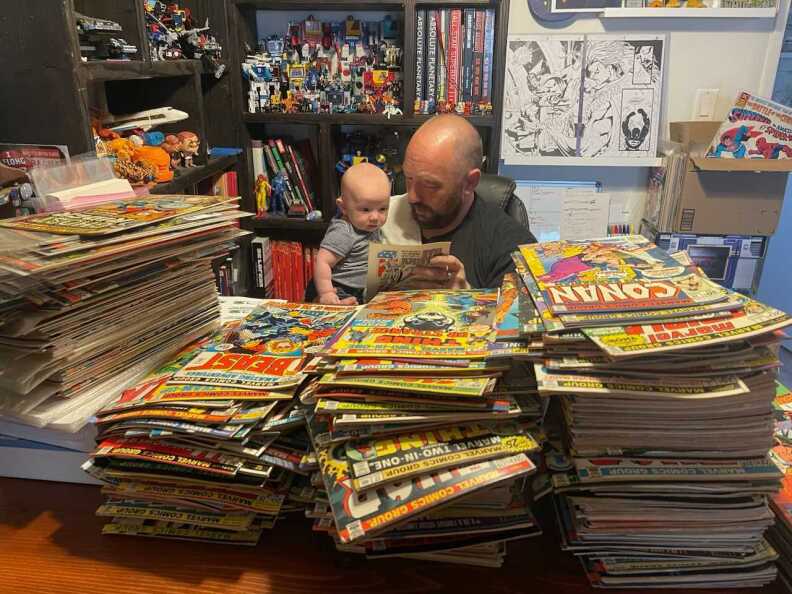 Geoffrey Patterson II and Geoffrey Patterson III sitting behind stacks of comic books as Patterson II holds a comic in front of Patterson III.