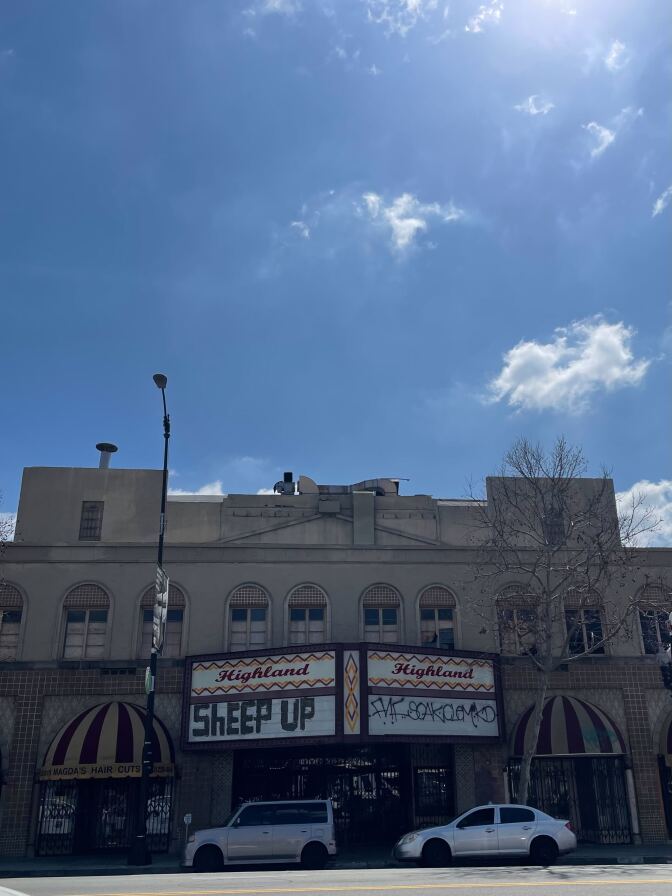 A view of Highland Theatre from across the street. A permanent sign reads "Highland" in red script, with a zig-zag border in red and yellow. On the white part of the sign where movie titles are usually posted, there is black graffiti text that appears to read "SHEEP UP."