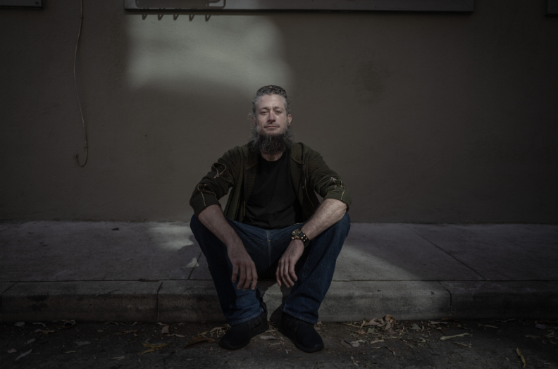 A white man with a long beard and greyish hair dressed in jeans and a greenish shirt over a black shirt is sitting down on a concrete sidewalk, looking at the camera.