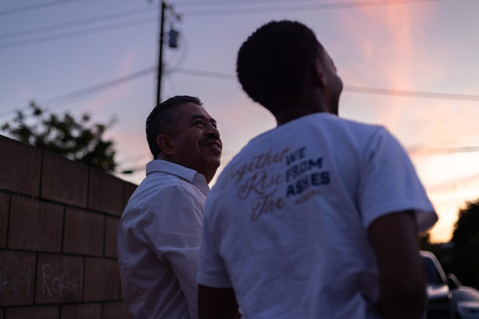Two men stand beside a hollow block fence in a backyard. The sky is colored orange, blue and grey.