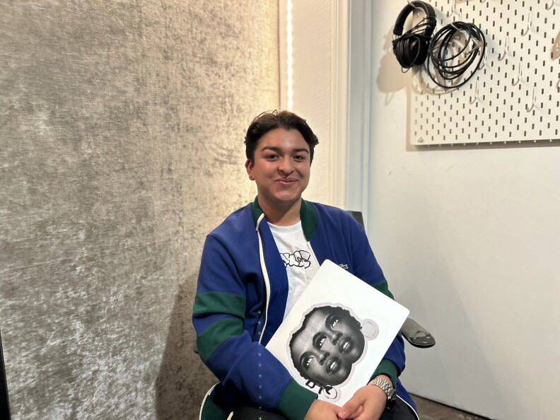 A young person with light brown skin, short dark hair smiles for the camera. They wear a blue sweater and hold a computer with a large sticker of a woman's face in black and white. 