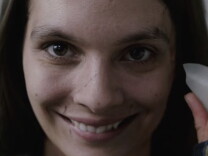 A photo of actress Caitlin Stasey in the horror movie "Smile." She is holding a sharp pottery shard to her face, which is scarred across her nose, and smiling in a disturbing manner.
