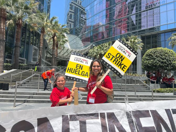 A woman with brown skin tone stands and smiles with her daughter while carrying picket signs.