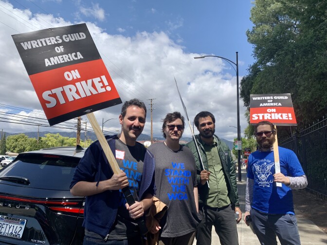 Four men stop from picketing to pose for a photo. Two carry signs that read "Writers Guild Of America On STRIKE!'