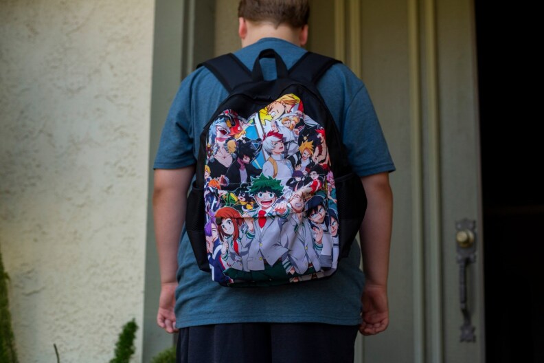 A student with his back to the camera wears a dark blue t-shirt and a backpack with anime characters.