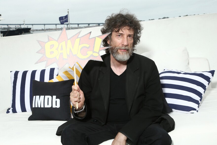 #IMDboat At San Diego Comic-Con 2018: Day Two with Neil Gaiman holding up a sign that says, 'Bang,' while sitting on a couch.