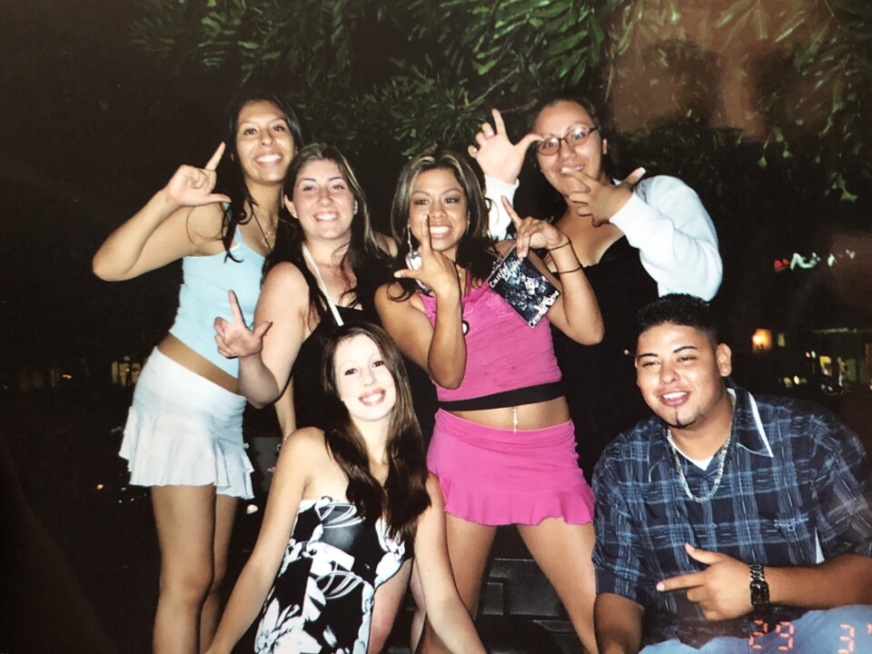 A diverse group of young people, mostly girls, pose for a photo in party clothes. All but one person are making an L gesture with their hands
