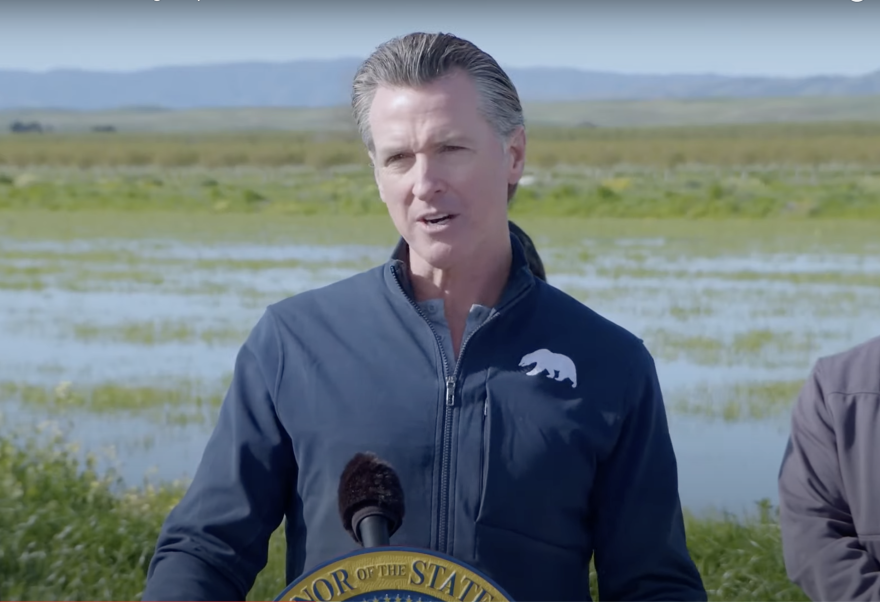 Governor Gavin Newsom stands in front of a podium emblezzoned with the blue and yellow California state seal. He stands with a federal blue jacket and slicked back grey hair. In the background are mountains and wetlands dotted with tall green grasses.