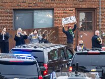 A small group of people, all with light-tone skin, stands along a red brick building as a motorcade of dark cars turns. A few people are waving and taking photos. One person holds a sign reading: "ABORTION IS NOT HEALTHCARE" and another holds a sign overhead that reads: "COMPLICIT in GENOCIDE."