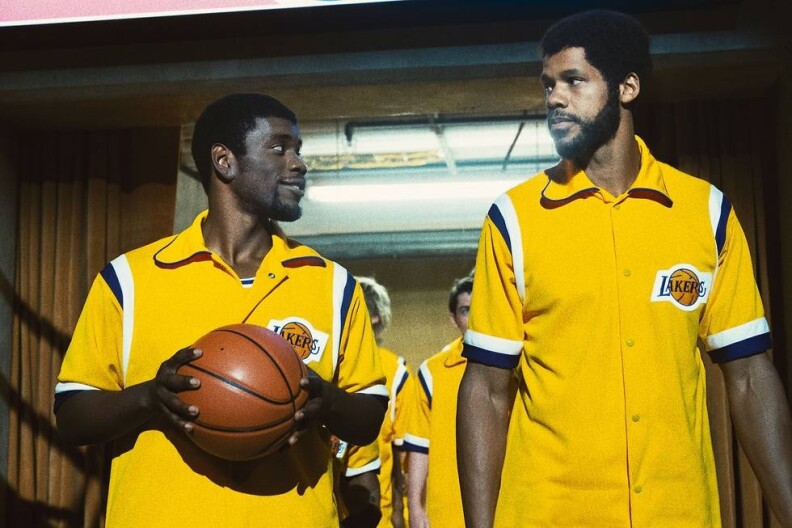 A man holding a basketball on the left, portraying player Magic Johnson, with another actor to the right, playing Kareem Abdul-Jabbar. Other men in Lakers pre-game uniforms are behind them. They each wear yellow button-up Lakers shirts as they walk out of a Forum tunnel.