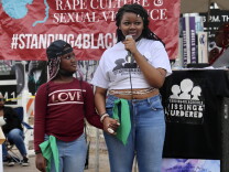 A young Black woman speaks into a microphone while holding the hand of a Black girl. The girl looks up at the woman, smiling. A banner is behind them that reads #standing4blackgirls. 