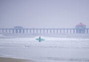 A surfer is walking through small waves near a sandy beach. A pier rises behind them in the water with cloudy skies beyond.