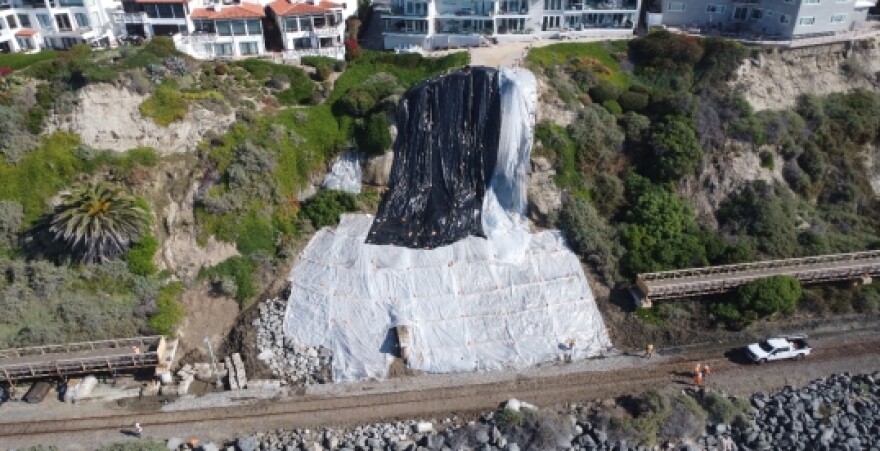 Black and white tarp cover a hillside where a landslide occurred. Beneath the hill is a train track that has been destroyed. A row of homes sit atop the hill.