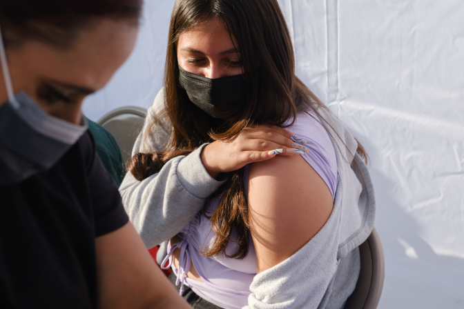 Noemi Zepeda, a 14-year-old, raises her sweater's sleeve as she awaits her COVID-19 vaccine. A health worker is seen in the foreground looking down.