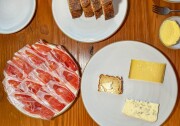A series of white ceramic plates are arranged on a medium-dark wooden tabletop. In the bottom left corner, overlapping slices of red and white cured ham cover one plate entirely. To the right, three small cheese slices display a range of colors: pale yellow, light mustard brown, and white, which is speckled with small black dots. Above this plate is a small dish containing a portion of yellow butter. At the very top, four slices of brown sourdough bread are positioned closely together on their sides.