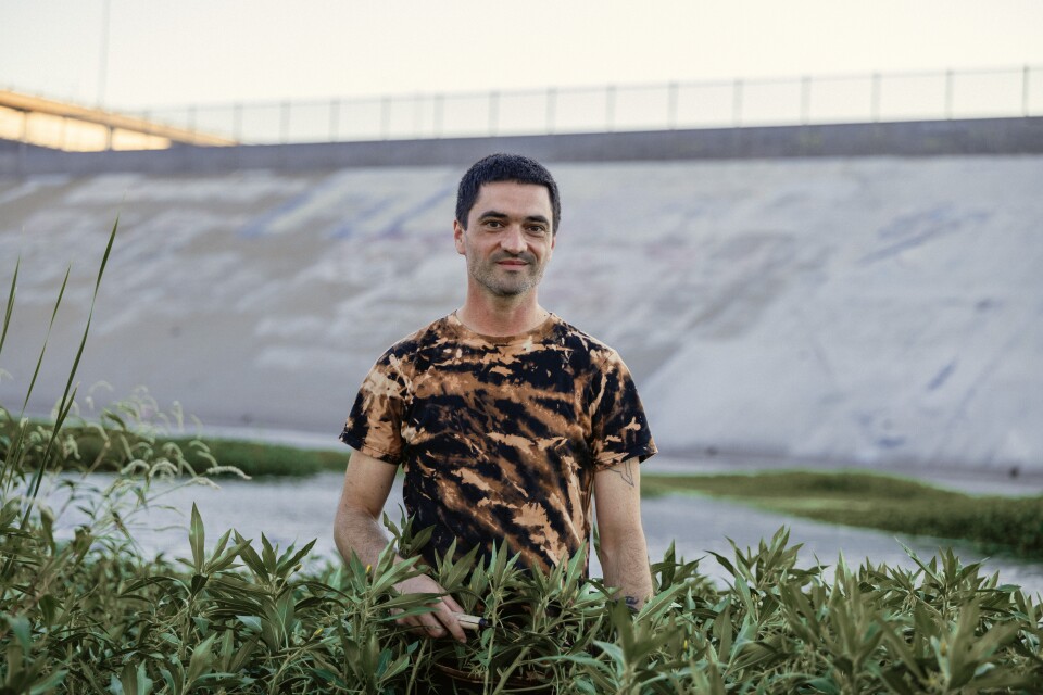 A man with medium skin tone and short hair, wearing a bleached-design t-shirt, stands behind greenery with the L.A. river and a concrete structure out of focus in the background.