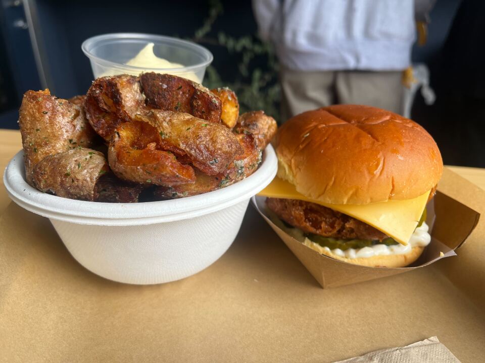 A paper tray on a counter top contains a fried fish sandwich that includes cheese, a fried fish patty, pickles, and mayo. Also on the tray is a bowl of crisp slices of roasted fingerling potatoes and a creamy dipping sauce.