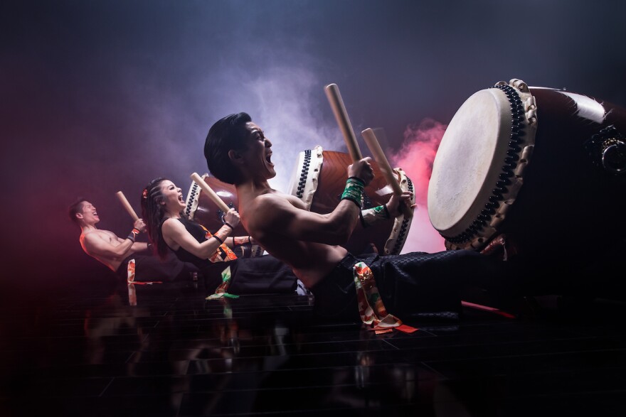 A line of three drummers play large taiko drums, with animated looks on their faces. Light seen through fog is behind them, with a darker background. There are men on either end, with a female taiko drummer in the middle.