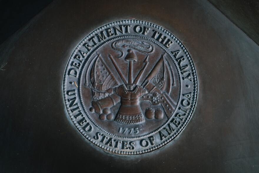 The emblem of the U.S. Army is depicted on a round medal with the words: Department Of The Army curved across the top and United States Of America along the bottom. In the middle is an engraving of various weapons and the date 1775.