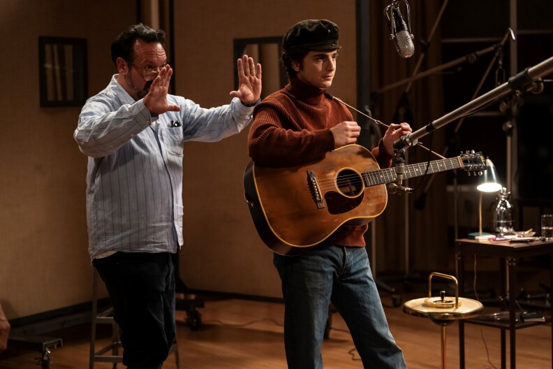 Actor Timothée Chalamet stands in costume as Bob Dylan at microphone, holding a guitar between takes for the 2024 film "A Complete Unknown." Behind him, director James Mangold is framing a shot with his hands.