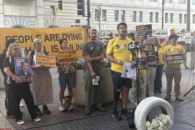 A person with a prosthetic leg speaks at a podium as others hold signs reading 'Traffic Violence Is A State Of Emergency' behind a 'People Are Dying' banner.