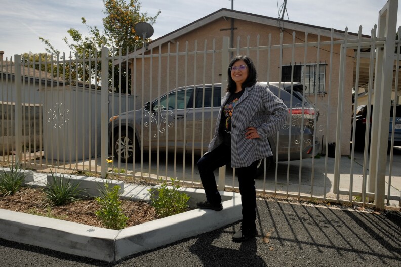 A middle-aged Latina woman smiles wearing a striped overcoat and black t-shirt and pants stands in front of a house with a metal fence and car parked in the driveway. She has one foot up on a curb that surrounds a mulched area with flowering plants.