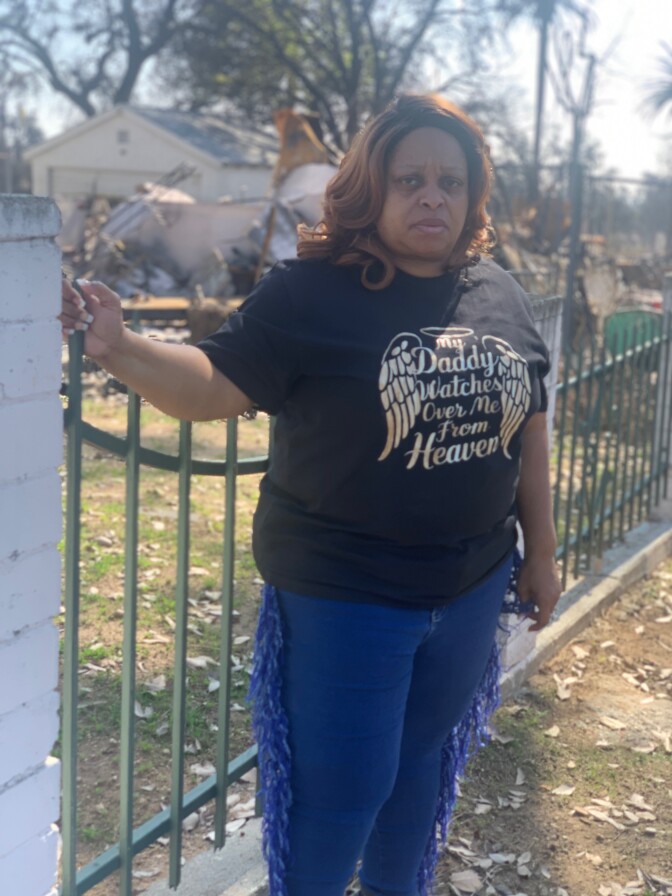 A middle-aged woman with dark brown skin stands in front of the rubble of a burned home. She wears jeans and a black t-shirt with gold lettering that reads "my Daddy watches over me from Heaven." 