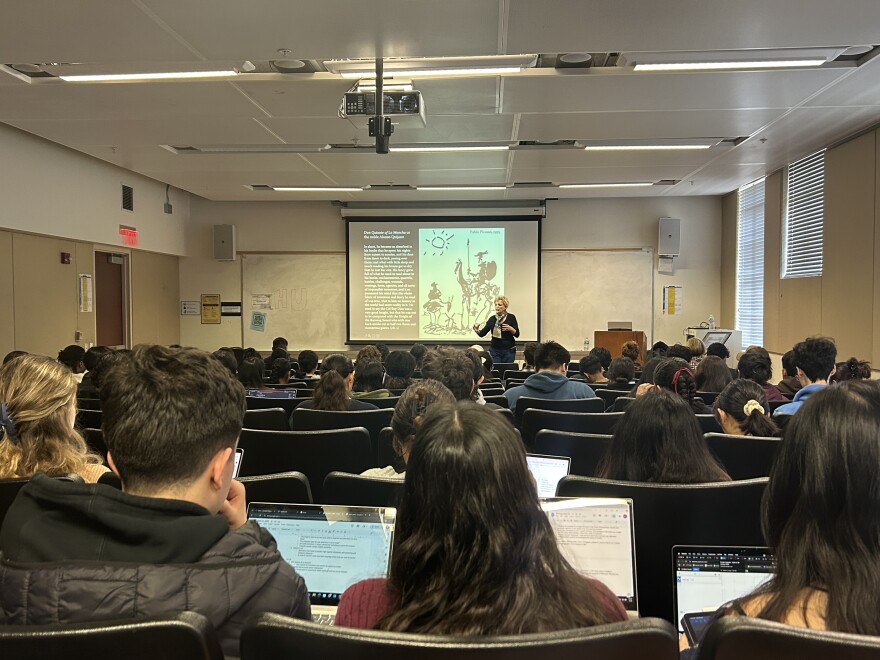 Rows of students with laptops face a woman with short hair in front of a projection screen. The screen features white text on a black background (illegible), along with Pablo Picasso's 1955 sketch of Don Quixote and and his sidekick, Sancho Panza.