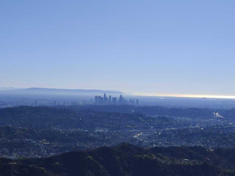 The metropolis of greater Los Angeles viewed from a mountaintop with the tall skyscrapers of downtown in the center and the sun gleaming off the ocean in the distance.