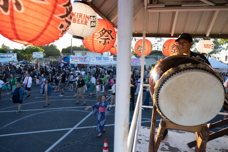 A view from inside the yagura, a platform raised above the crowd. An Asian American man with a baseball cap plays a taiko drum as a crowd of people dance below.
