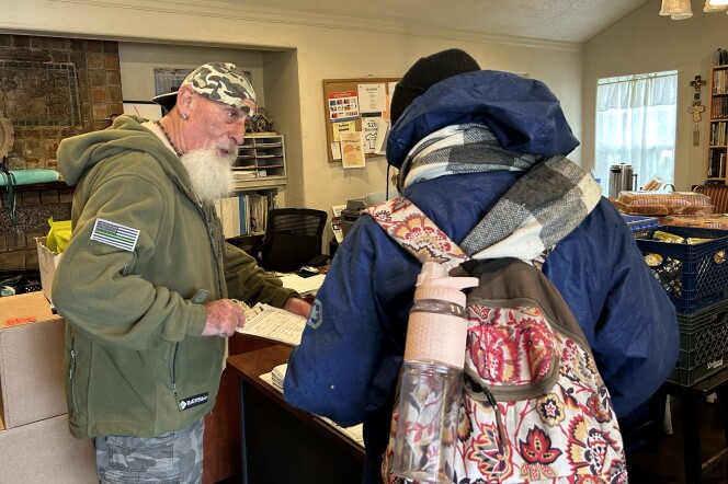 A man in an office with a full, white beard wearing an Army green sweatshirt holds a clipboard with a form on it out to another man, seen from the back, wearing a blue hooded jacket and carrying a flower print backpack. 