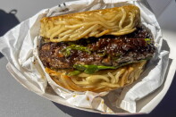 A close-up of a ramen burger — a beef patty glazed in dark sauce with arugula, sandwiched between two seared noodle buns — resting in white food paper.