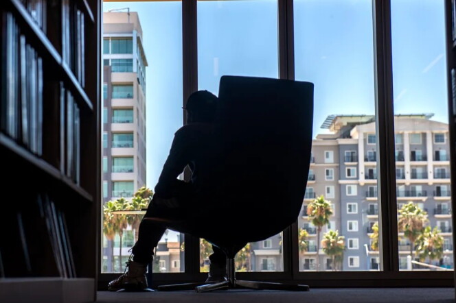 A man sits in one of the oversized chairs next to shelves of books inside a library.