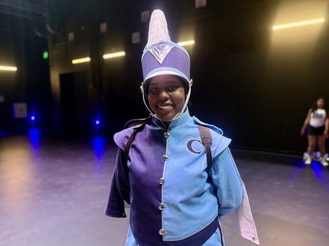 A student with dark brown skin tone in blue marching band attire smiles for a photograph.