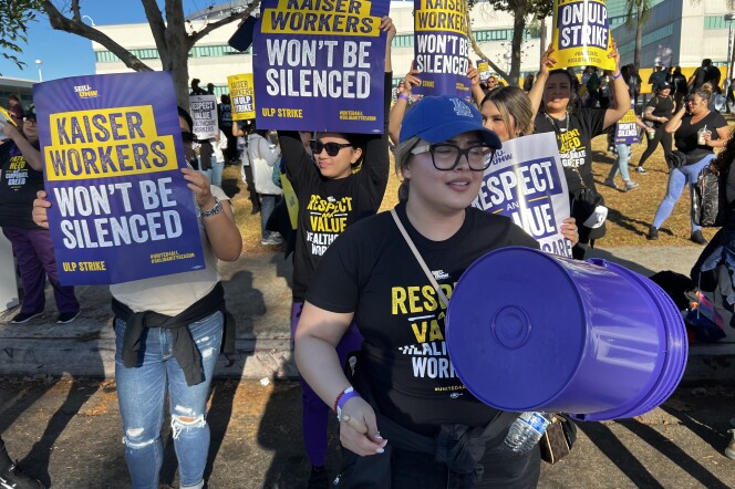 A crowd of people picket outside a hospital. They're wearing black union shirts while holding signs that read "Kaiser workers won't be silenced."