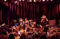 Dwight Yoakam plays to a crowd at The Roxy. He plays a acoustic guitar and wears a blue jacket and white cowboy hat. 