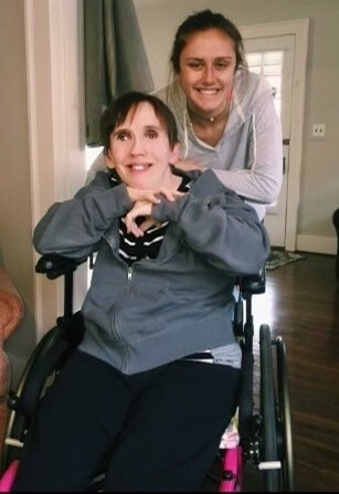 Kristy Barrett sits in a wheelchair with her niece Kendall Barrett standing behind her. Both are smiling.
