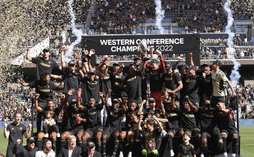 Two dozen men with a range of skin tones wearing black shirts and hats with gold lettering stand with their arms in the air on a platform in the middle of a green soccer field in front of a banner that reads "Western Conference Champions 2022" in front of a row of stands filled with fans in the background at Banc of California Stadium in South L.A. One man in the center holds a silver cup trophy above his head as the other men cheer.