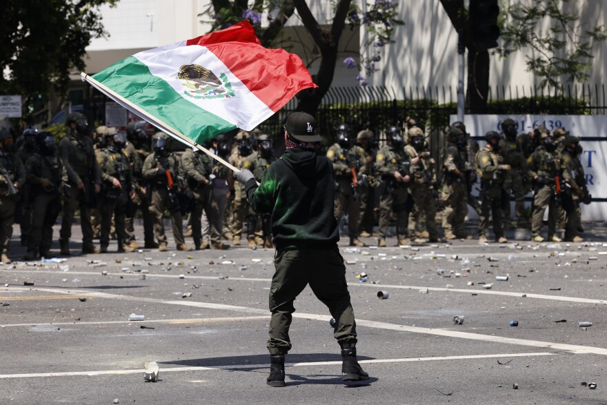 A man standing in a street waves a Mexican flag. He's wearing a black hat, hoodie, dark green pants and boots. People in military gear stand in the distance.