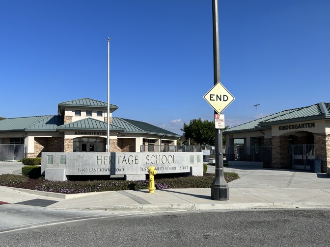 A wide shot of a school with a large concrete sign in front reading Heritage School. To the right, there's a building with a green roof labeled Kindergarten. To the left is a building with a green roof labled Administration. 