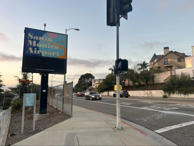 Signage that reads "Santa Monica Airport" stands at a street intersection with cars driving by and homes and apartments across the street.