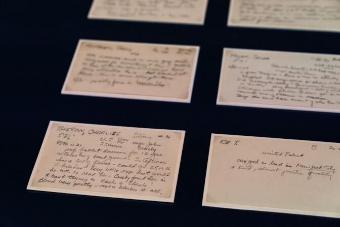 A white 3 by 5 index card in a museum gallery case, in a row with several other cards on a black background, with casting director Marion Dougherty's hand-written notes about her impressions of Charlize Theron. She describes her as “blond very pretty — not a bimbo at all” and says she “has a lot of poise.”


