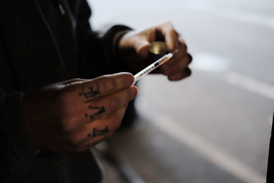 A person in a dark sweater holds a skinny syringe with a light brown liquid inside. In the other hand is a small shiny gold cap.