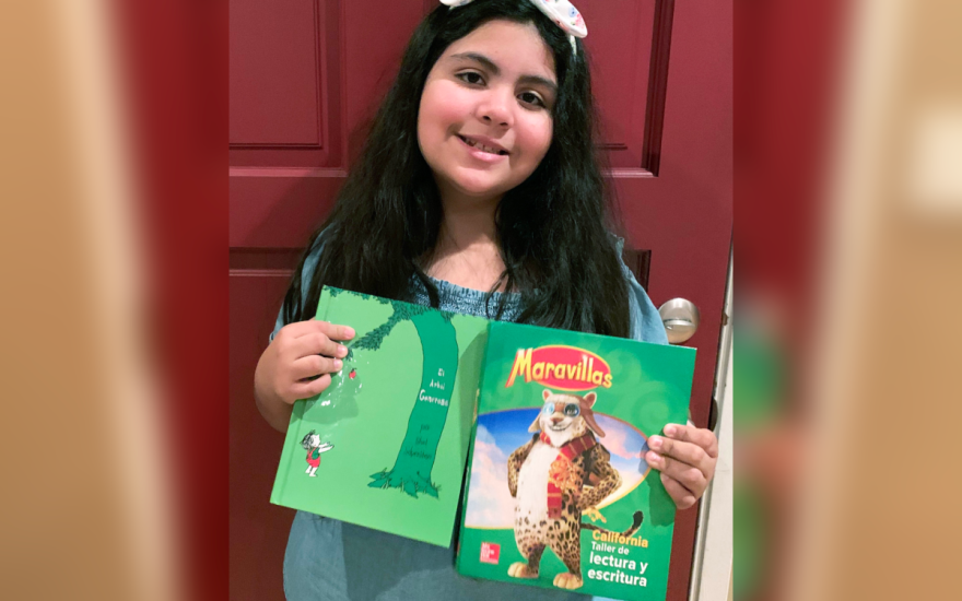 A young girl holds a Spanish-language book with a picture of a tiger on the front. The book's title is "Maravillas: California Taller de lectura y escritura."